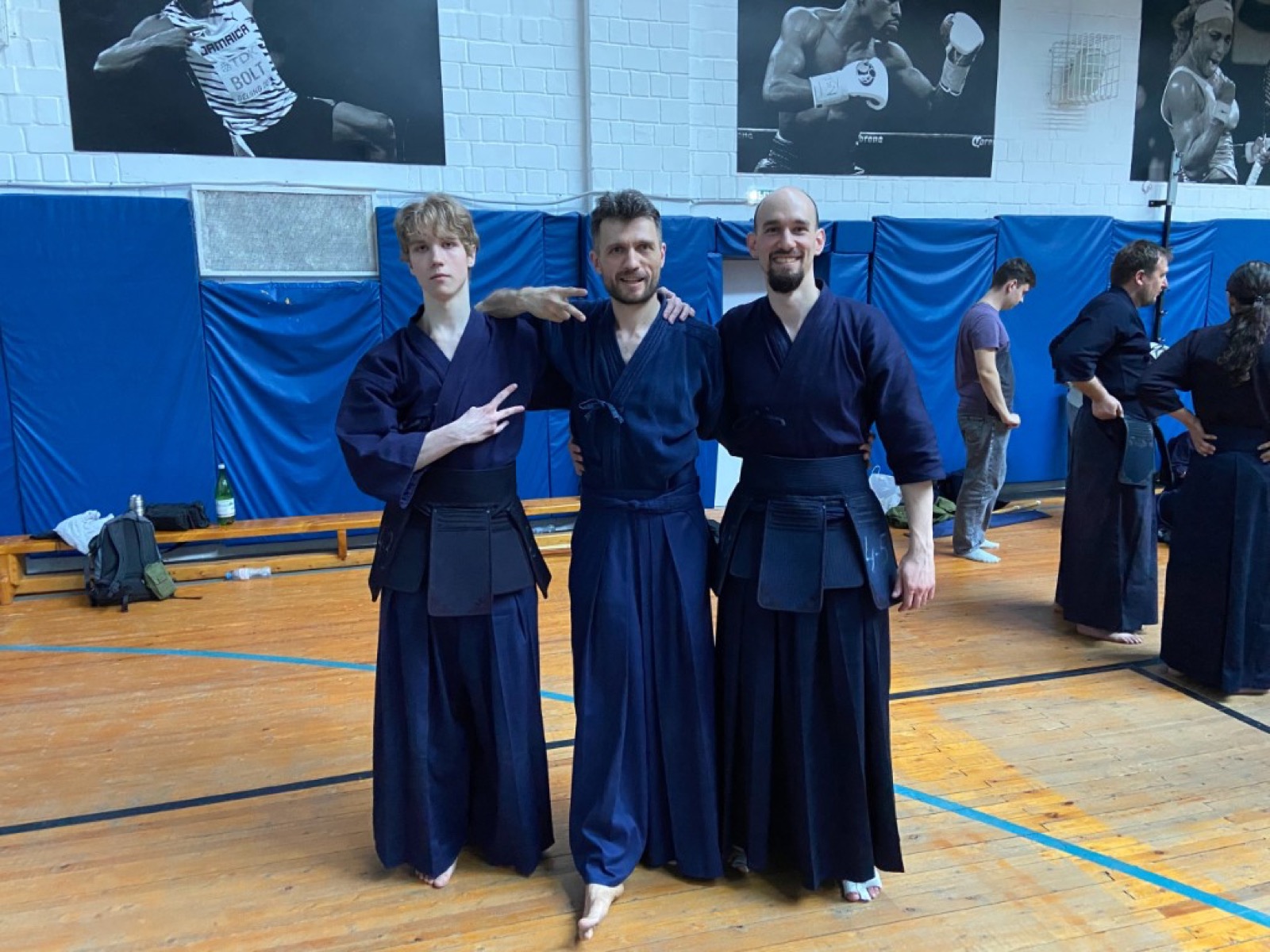 Трое кендоков в тёмно-синей форме улыбаются в камеру в спортзале, средний показывает знак мира